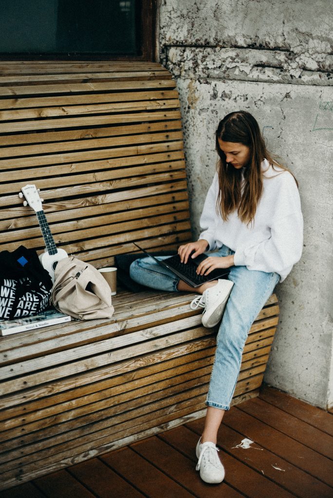 Kobieta w białej koszuli, dżinsach i białych trampkach siedzi z laptopem na nodze. Oparta jest o szary mur. Naprzeciwko niej na ławce leżą torby, książka, gitara i papierowy kubek.