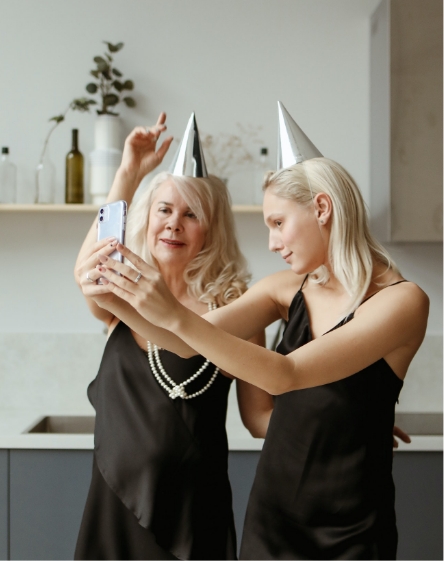 Dwie kobiety w czarnych sukienkach, o blond włosach i srebrnych czapkach urodzinowych na głowach. Jedna z nich trzyma telefon i robi zdjęcie selfie. W tle widać półkę z wazonami i szare szafki.