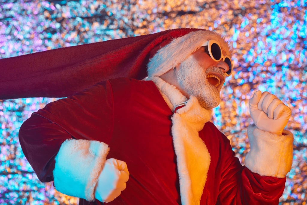 Biegnący mężczyzna w przebraniu Świętego Mikołaja z białymi okularami i otwartymi ustami na kolorowym tle.