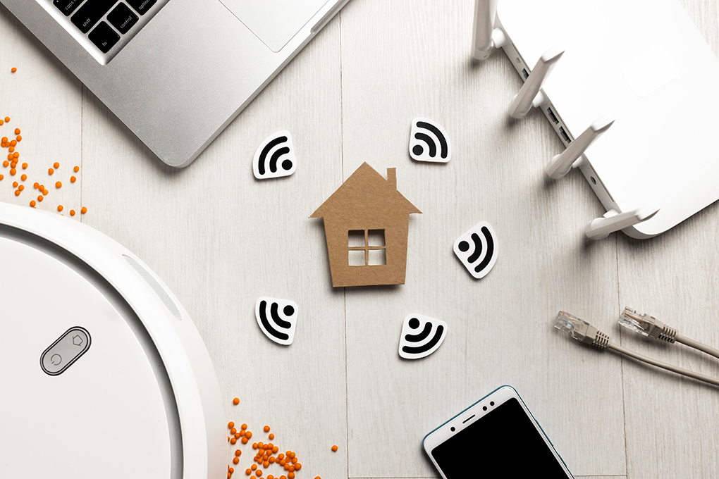 Na środku obrazka ikonka domu otoczona znaczkami wifi. Naokoło tej ikonki router, kabel, telefon, laptop, odkurzacz