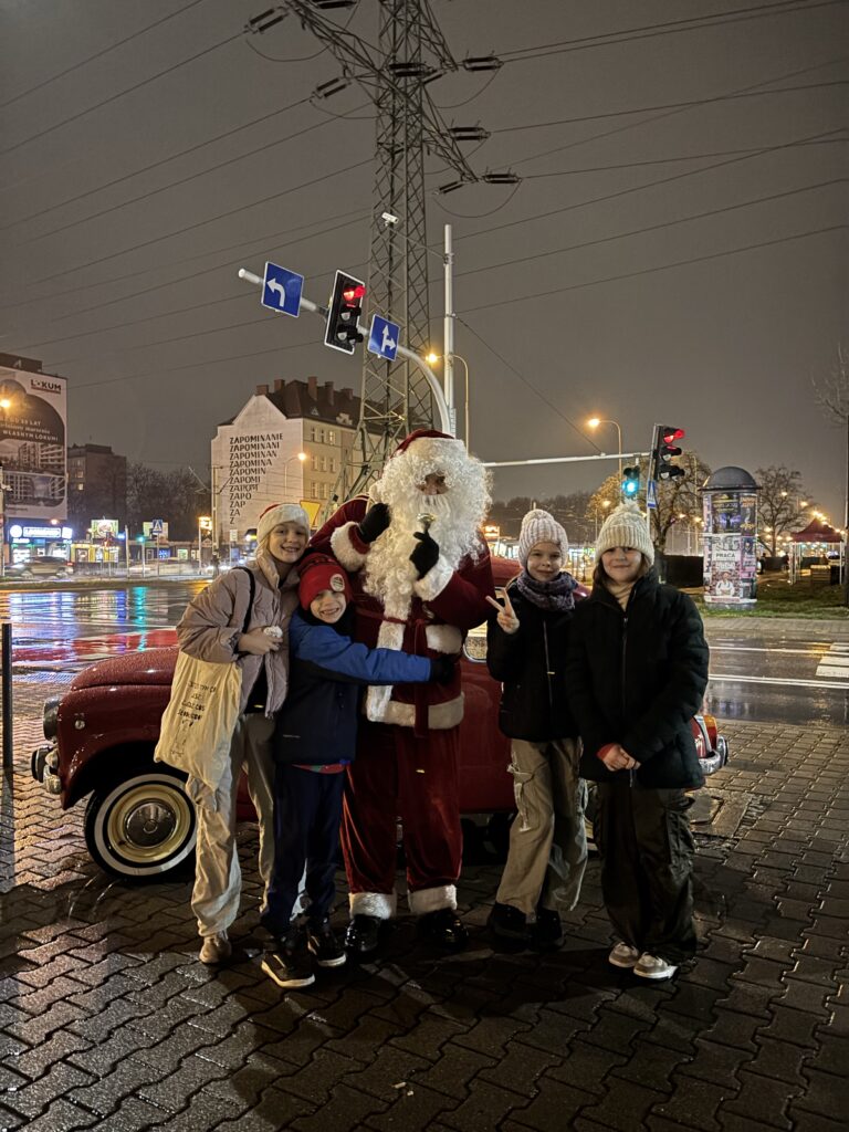 Na zdjęciu widzimy grupę osób pozujących z MOICOłajem w zimowej scenerii miejskiej. MOICOłaj, ubrany w tradycyjny czerwony strój z białym futerkiem, stoi obok zabytkowego samochodu, który dodaje nostalgicznego klimatu. Otaczają go cztery osoby ubrane w zimowe ubrania, takie jak kurtki, czapki i szaliki, co podkreśla zimową aurę. W tle widać oświetlone miasto z ulicznymi latarniami, znakami drogowymi oraz elementami miejskiej infrastruktury