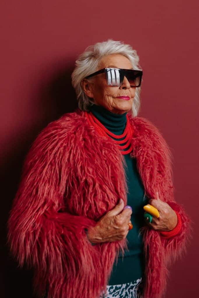 Na zdjęciu widzimy starszą kobietę, która emanuje pewnością siebie i wyrazistym stylem. Ubrana jest w intensywnie czerwoną, futrzaną kurtkę, pod którą znajduje się ciemnozielony golf. Stylizację uzupełniają liczne czerwone korale, bransoletka oraz duże, lustrzane okulary przeciwsłoneczne, a na palcach duże, kolorowe pierścionki.