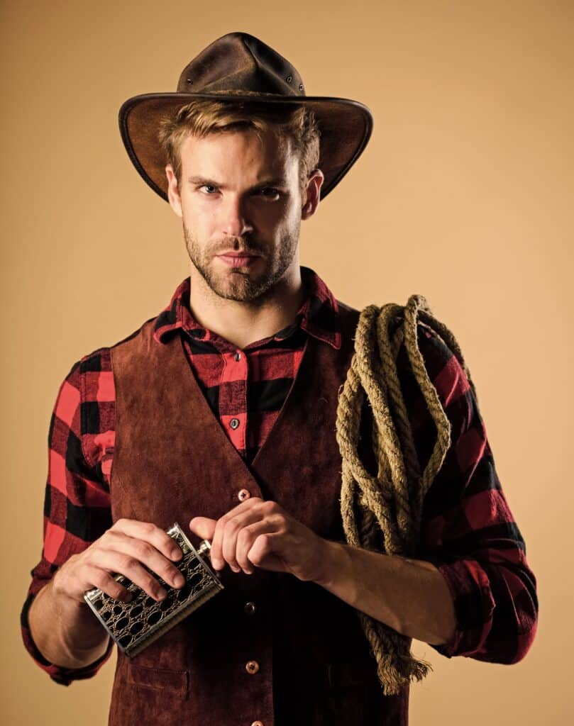 Na zdjęciu widzimy mężczyznę ubranego w styl kowbojski, który z dumą prezentuje klasyczne elementy amerykańskiego Dzikiego Zachodu. Na głowie ma brązowy skórzany kapelusz, a ubranie składa się z czerwono-czarnej koszuli w kratę oraz brązowej zamszowej kamizelki. Przez ramię przewieszony ma gruby, zwinięty sznur, a w dłoni trzyma niewielki metalowy przedmiot o kratkowanej strukturze, w którym odkręca korek.