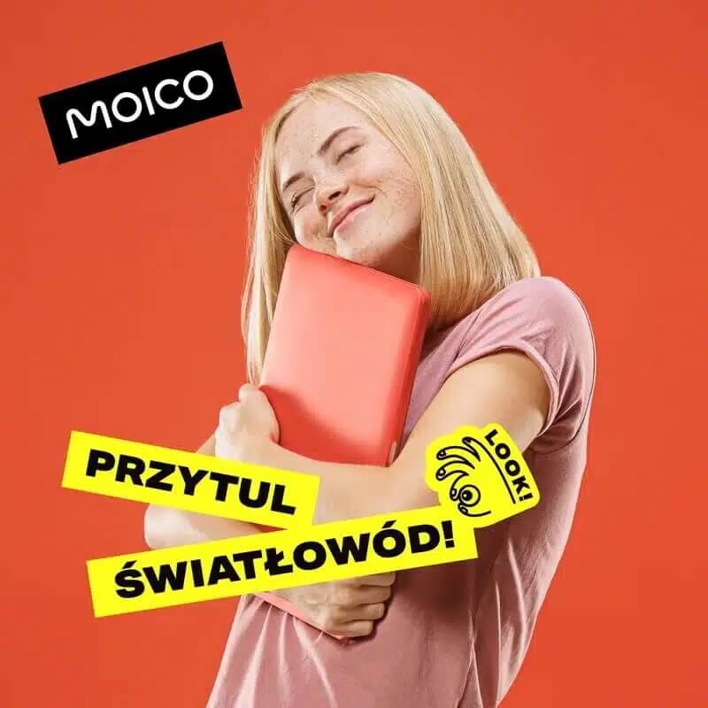 Na pomarańczowym tle widzimy kobietę w różowej koszulce, która z radością przytula czerwony prostokątny obiekt. Hasło „PRZYTUL ŚWIATŁOWÓD!” w żółtym kolorze zachęca do emocjonalnego związania się z technologią. Logo „MOICO” wskazuje na markę promującą usługę światłowodową. Dodatkowy element graficzny z napisem „LOOK!” i okiem dodaje kampanii energii i młodzieżowego charakteru.