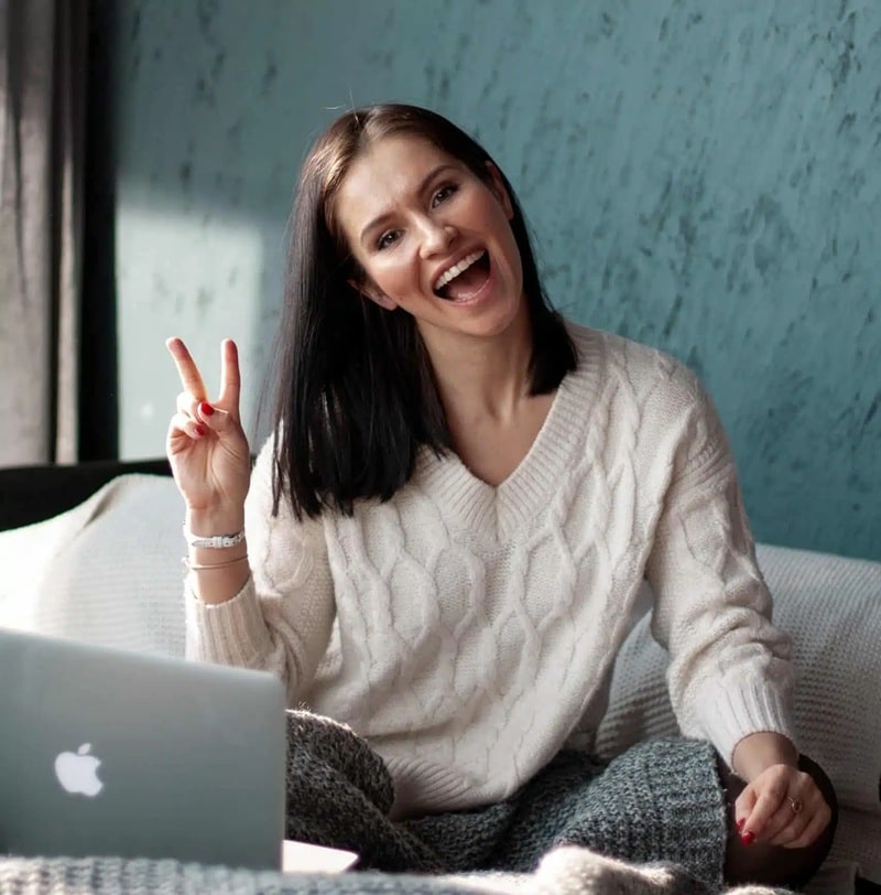 Uśmiechnięta kobieta w białym swetrze i szarych spodniach siedzi wygodnie przed otwartym laptopem, pokazując znak pokoju. Naturalne światło i turkusowe tło tworzą przytulną, domową atmosferę. 