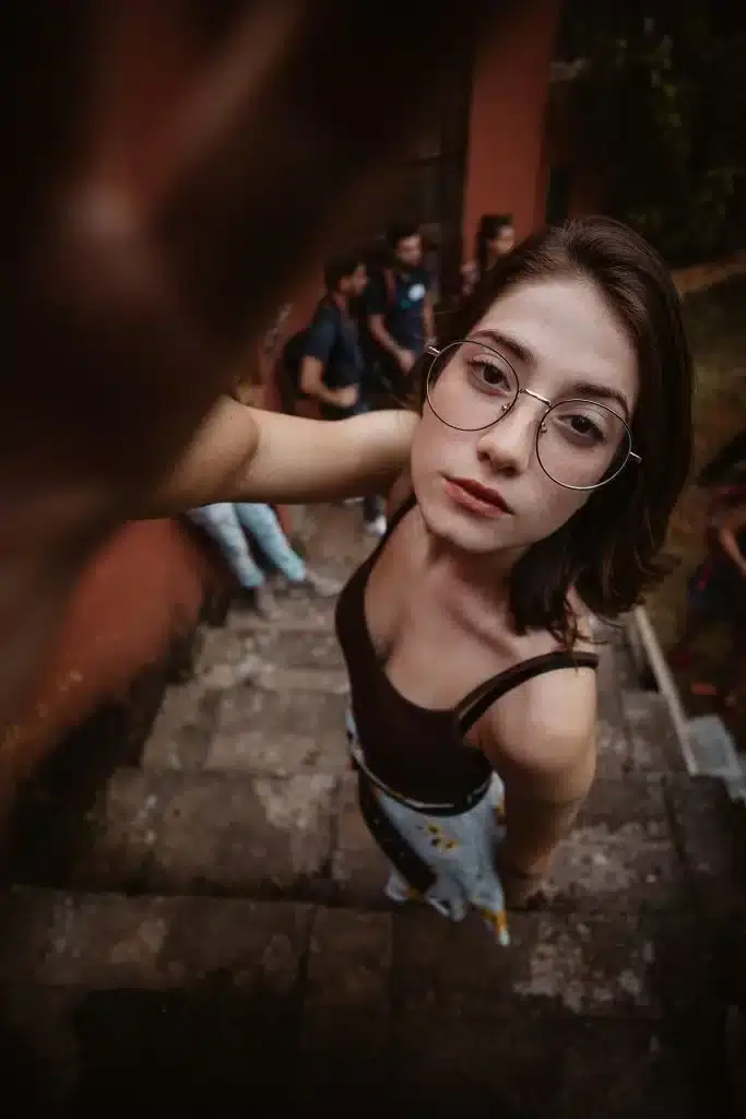 Na zdjęciu przedstawiona młoda kobieta, która stoi na kamiennych schodach, patrząc prosto w obiektyw z neutralnym wyrazem twarzy. Ujęcie z góry podkreśla głębię kompozycji, a duże okulary i wzorzyste spodnie nadają stylizacji charakteru. W tle widać innych ludzi oraz otoczenie z zielenią i ceglanym budynkiem, co tworzy swobodną, miejską atmosferę.