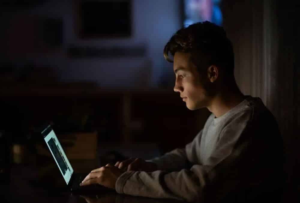 Na zdjęciu widać młodego mężczyznę siedzącego przy laptopie w słabo oświetlonym pomieszczeniu. Mężczyzna jest skupiony na ekranie, który oświetla jego twarz. Ma na sobie szarą bluzę.