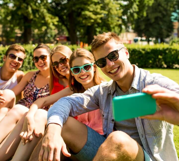 Na zdjęciu widać grupę pięciu młodych osób siedzących razem na trawie w słoneczny dzień, prawdopodobnie w parku. Jedna z osób trzyma telefon i robi wspólne selfie, a pozostali uśmiechają się do obiektywu. Wszyscy mają na sobie letnie ubrania i okulary przeciwsłoneczne, co podkreśla wakacyjny, swobodny klimat.