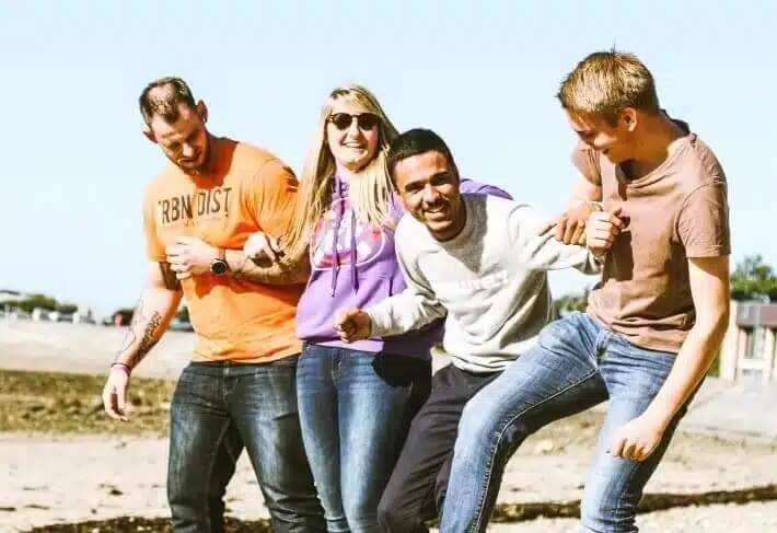 Na zdjęciu widać czworo młodych ludzi spędzających razem czas na świeżym powietrzu. Stoją blisko siebie, śmieją się i wygłupiają, co sugeruje swobodną, przyjacielską atmosferę. Ubrani są casualowo – w jeansy, bluzy i T-shirty. Jedna z osób ma okulary przeciwsłoneczne.