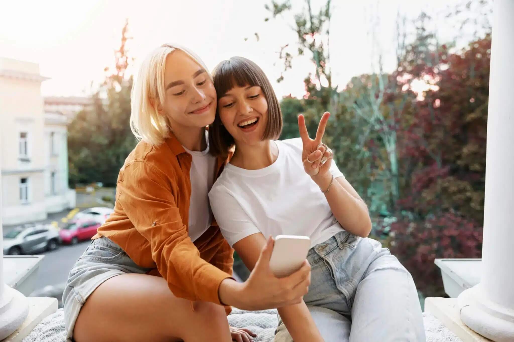 Na zdjęciu znajdują się dwie młode kobiety siedzące obok siebie na tarasie lub balkonie. Jedna z nich trzyma smartfon i robi wspólne selfie, a druga pokazuje dłonią znak pokoju. Obie są uśmiechnięte i wyglądają na zadowolone, a w tle widać drzewa, budynki oraz zachodzące słońce.