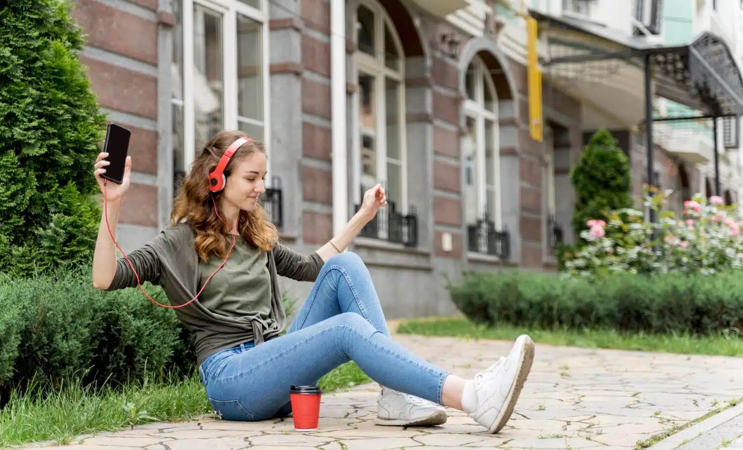 Na zdjęciu widać młodą kobietę siedzącą na chodniku przed budynkiem mieszkalnym. Ma na uszach czerwone słuchawki przewodowe podłączone do smartfona, który trzyma w dłoni. Kobieta uśmiecha się i wygląda na zrelaksowaną, słuchając muzyki. Obok niej stoi czerwony kubek z napojem. W tle znajdują się zadbane krzewy oraz zabudowa wielorodzinna, co sugeruje miejskie osiedle.