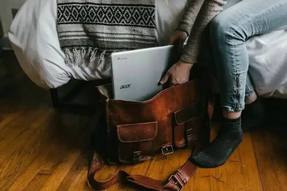 Na zdjęciu widać osobę siedzącą na łóżku i pakującą laptopa do dużej, skórzanej torby. Laptop znajduje się w centralnej części kadru, a obok widoczny jest szeroki pasek torby. Osoba ma na sobie sweter, jeansy i ciemne skarpetki.