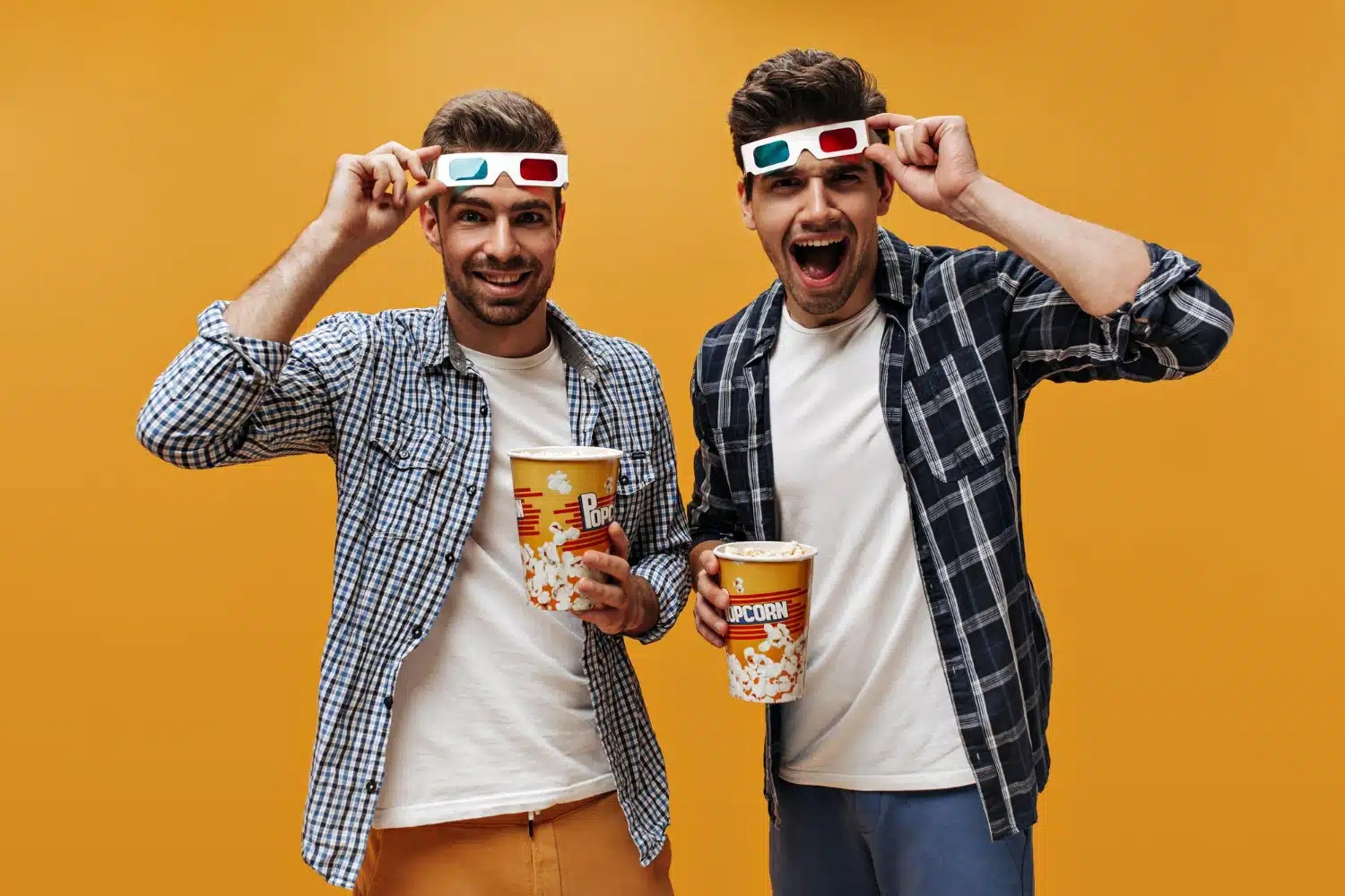 Na zdjęciu widać dwóch mężczyzn ubranych w casualowe koszule w kratę i białe T-shirty. Obaj trzymają kubki z popcornem i mają na sobie okulary 3D. Mężczyźni uśmiechają się i wyglądają na podekscytowanych, a całość przedstawiona jest na jednolitym, pomarańczowym tle.