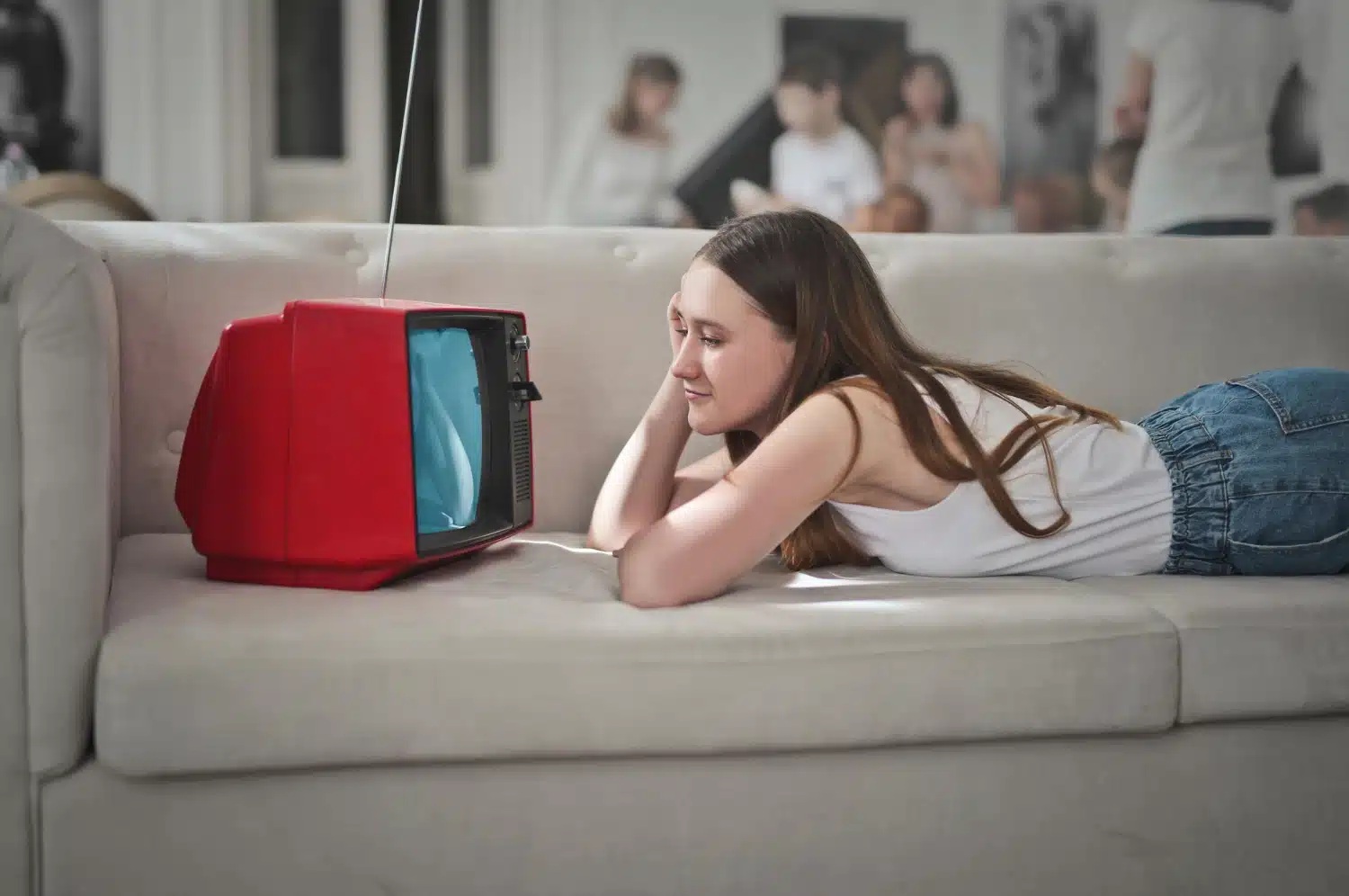 Na zdjęciu widać kobietę leżącą na jasnej kanapie i oglądającą telewizję. Obok niej znajduje się niewielki, czerwony telewizor w stylu retro. W tle widać odbicie kilku osób w lustrze, a cała scena przedstawia domowy, swobodny sposób spędzania czasu przed telewizorem.